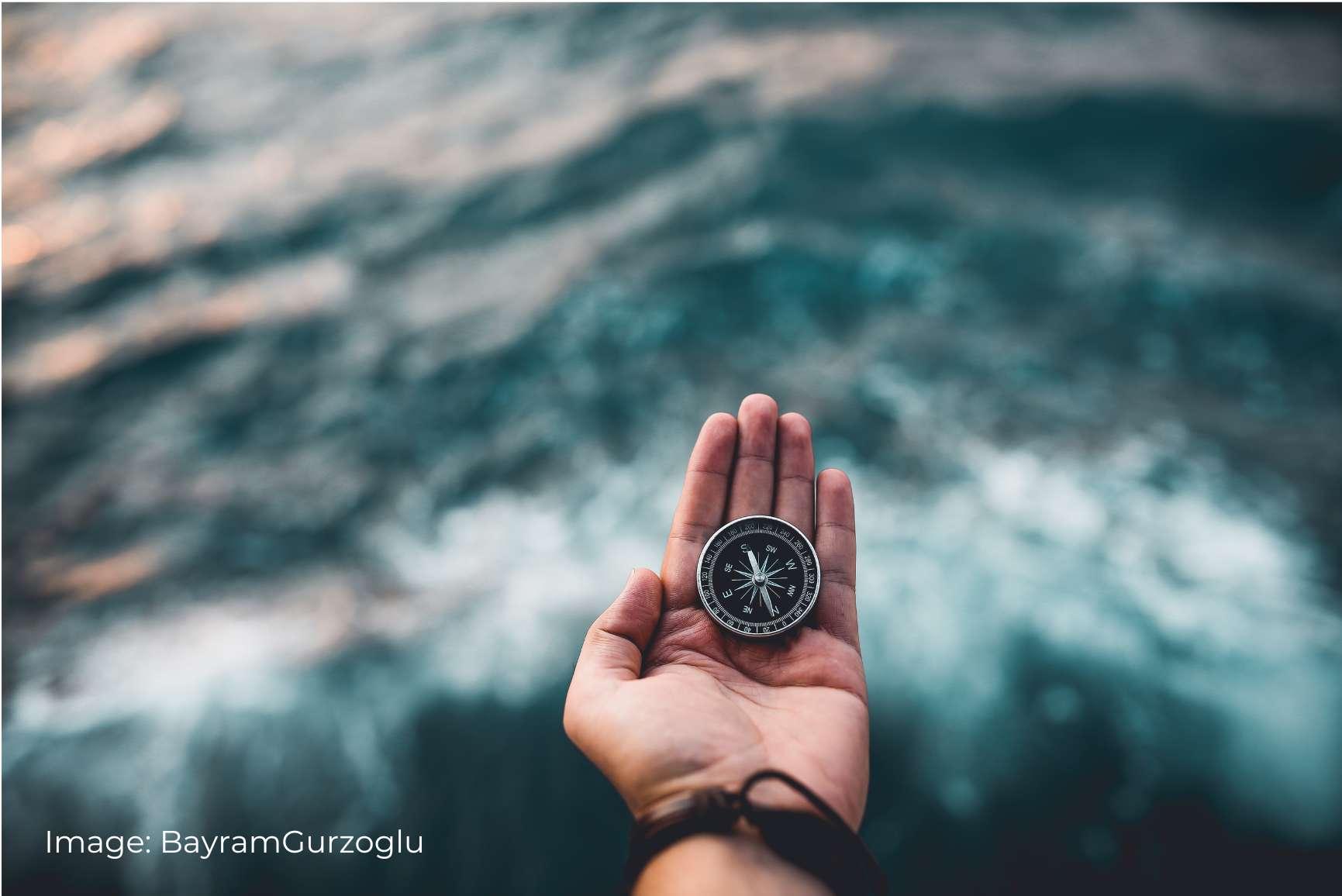 A hand holding a compass over stormy water, symbolizing the ability to navigate through challenging conditions. The blurred background of ocean waves enhances the feeling of exploration and resilience. Image by BayramGurzoglu from Pexels