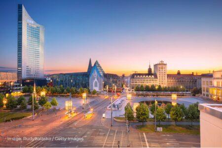 Panoramic view of Leipzig at sunset, showcasing the modern Panorama Tower where the EEX headquarters is located. The image also features the distinctive Paulinum building of the University of Leipzig, surrounded by well-lit streets, green trees, and historic architecture. The vibrant urban landscape is captured beautifully with a mix of modern and historical elements. Image by RudyBalasko from Getty Images.